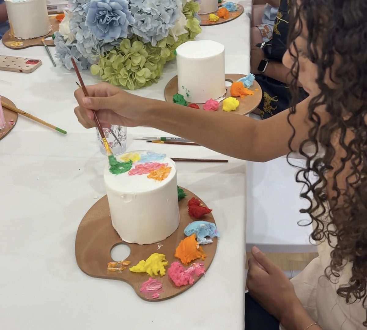 A girl painting coloured buttercream onto a white cake from an artist’s palette