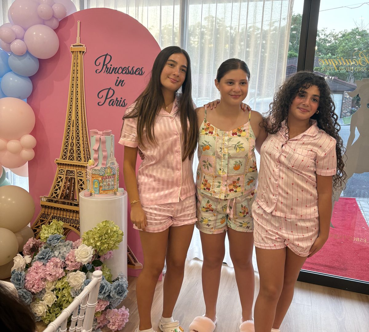 Three teenage girls in pyjamas in front of a Princesses in Paris balloon backdrop