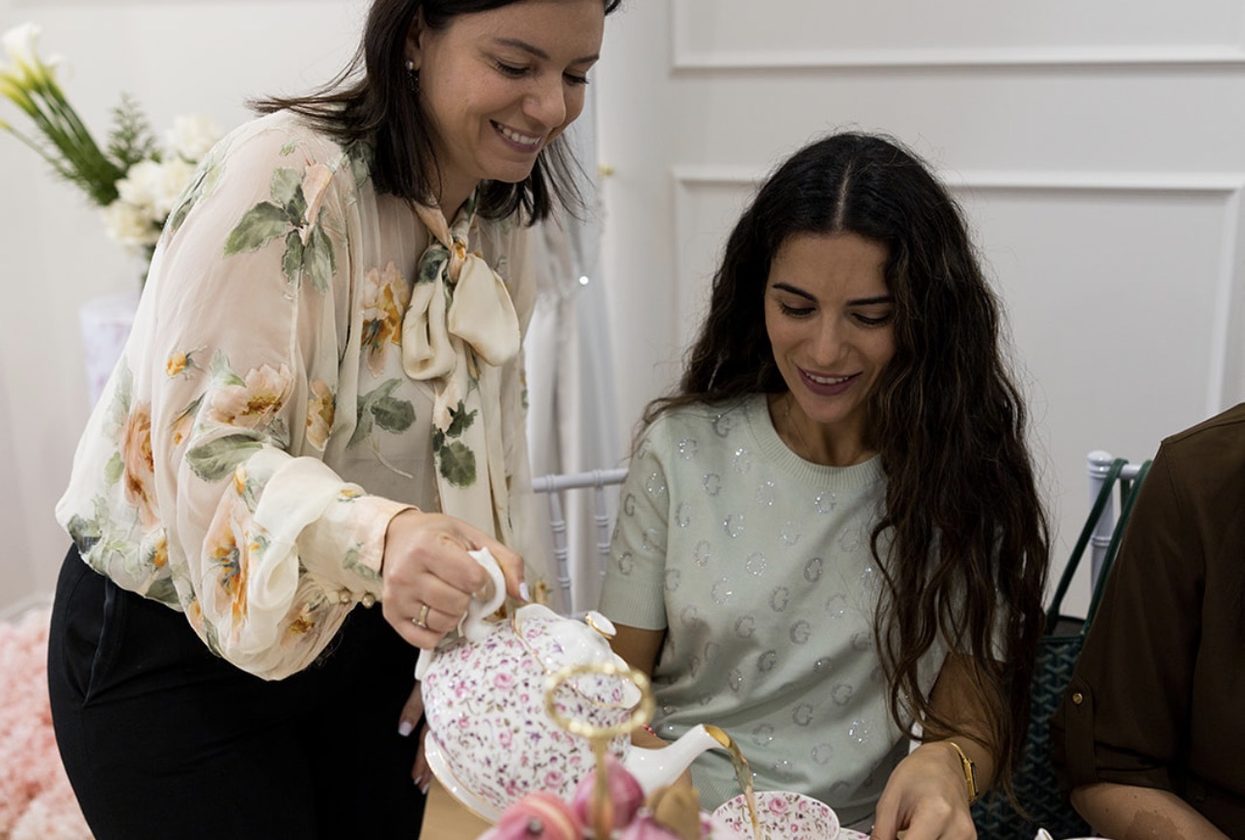 A mum pouring tea for a seated guest beside a tiered high tea stand