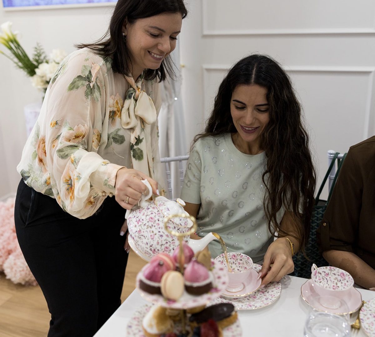A mum pouring tea for a seated guest beside a tiered high tea stand
